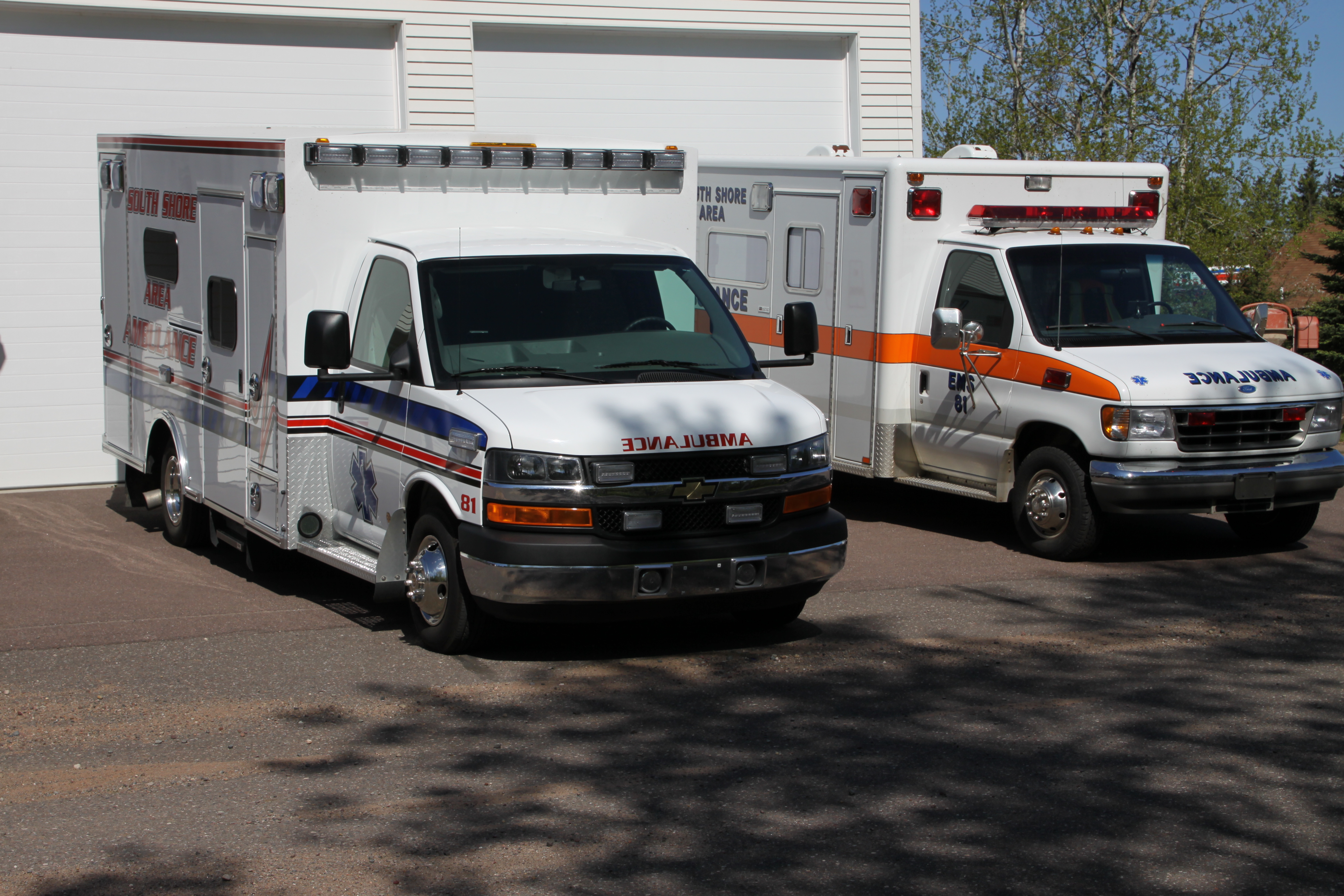 Passenger side view of the two Bayfield County South Shore Ambulances.  One with red and blue stripes the other white with a ora